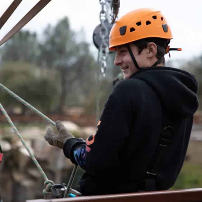 A young person in an orange helmet and gloves participates in an outdoor rock climbing course, smiling while secured with safety harnesses.