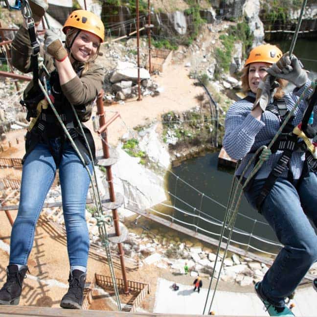 Two people rappelling over an abandoned industrial site, wearing helmets and harnesses, smiling and looking excited.