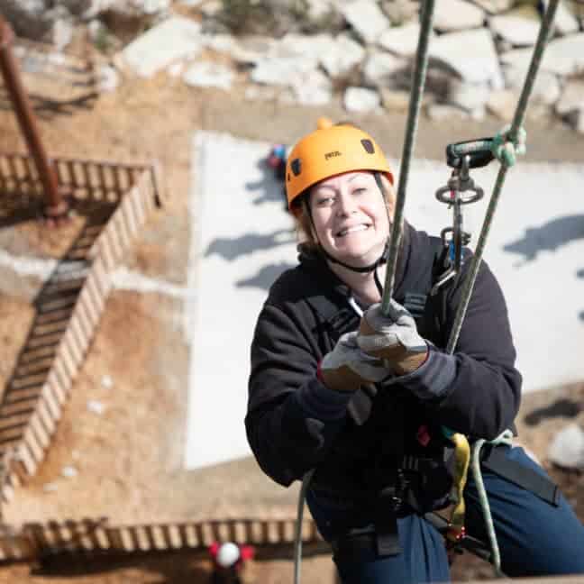 A woman in climbing gear and gloves smiles while zip lining over a stone courtyard with other participants below.