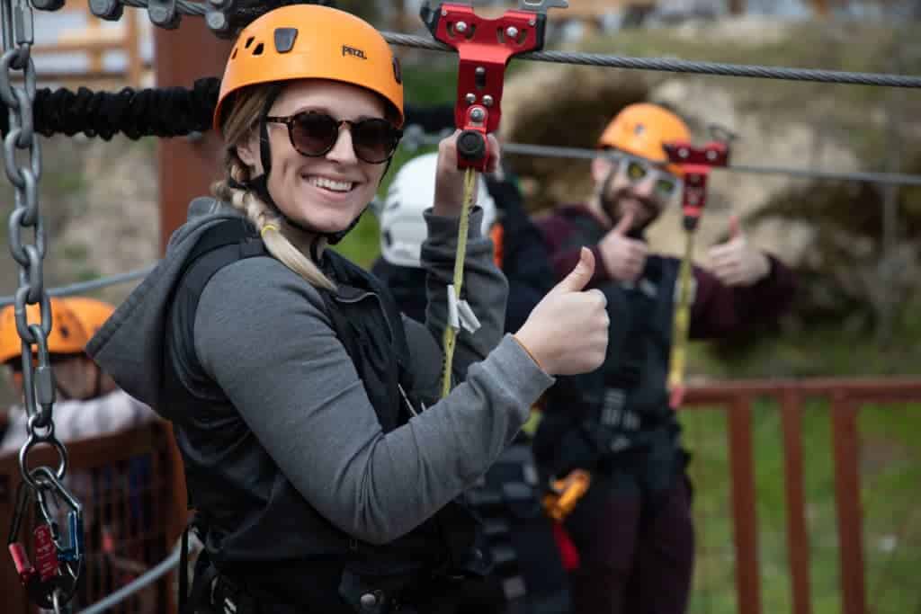 Woman wearing a helmet smiles and gives a thumbs-up during a corporate team building event; man in the background also thumbs-up, both secured with zipline safety gear.