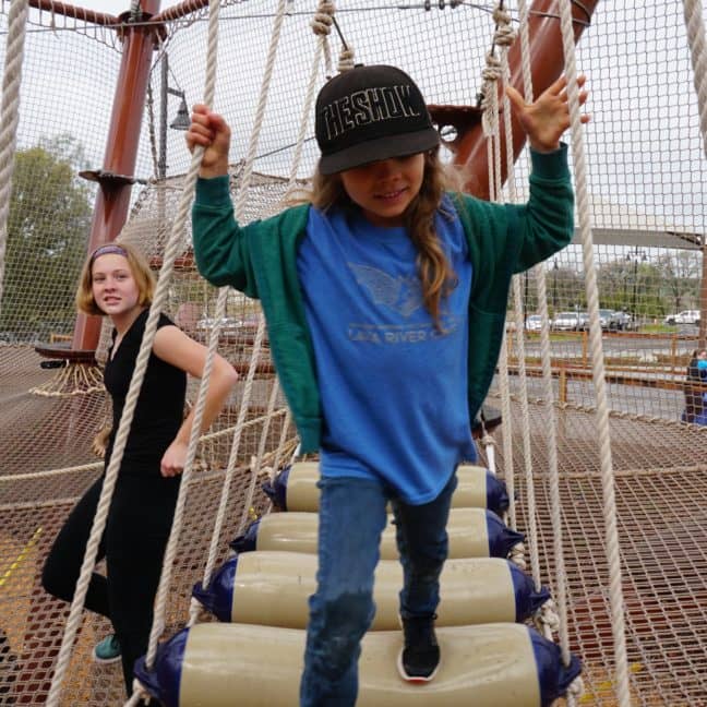 A young girl in a blue sweatshirt navigates a rope bridge obstacle at a playground with other children in the background.