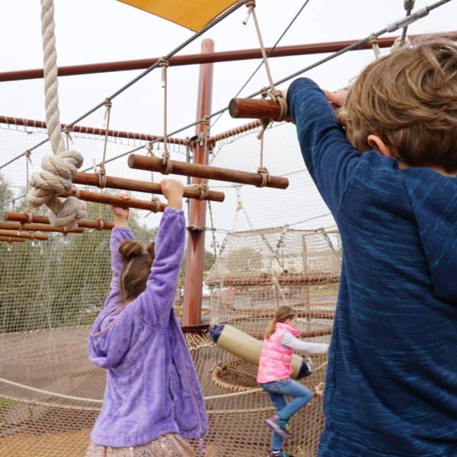 Children play on a rope and wooden ladder structure at a playground, with one boy in focus climbing towards a suspended platform.