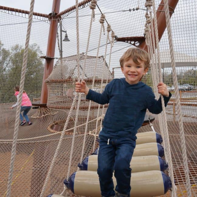 A young boy smiles while playing on a rope bridge at a playground, with other children in the background.