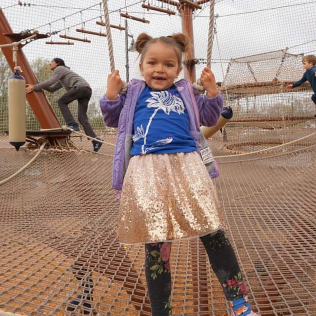 Toddler girl smiling on a rope swing at a playground, wearing a purple jacket and sparkly skirt.