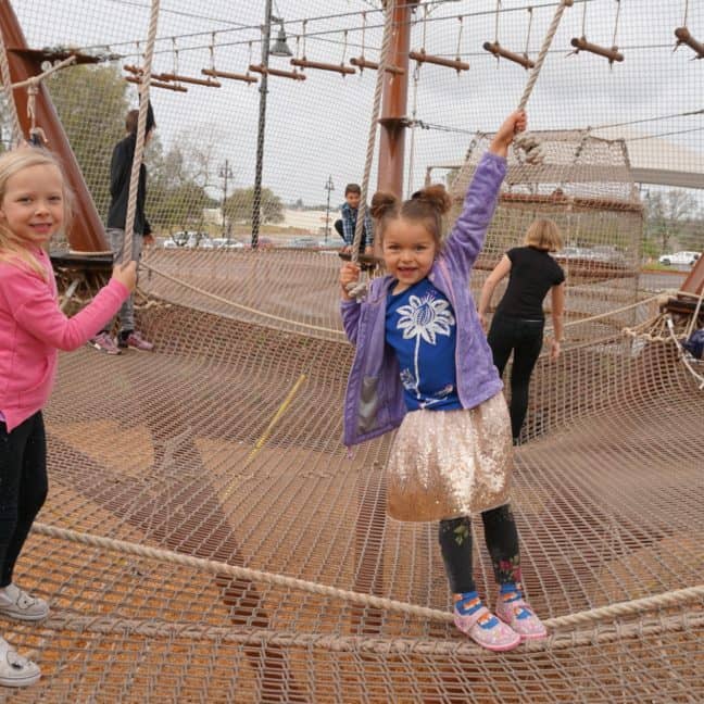 Two young girls playing on a netted climbing structure with suspended punching bags at a playground.