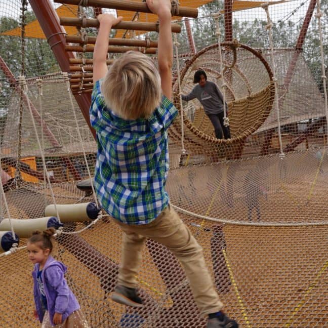 A child in a plaid shirt swings from a horizontal rope in a busy playground, while another child in purple watches.