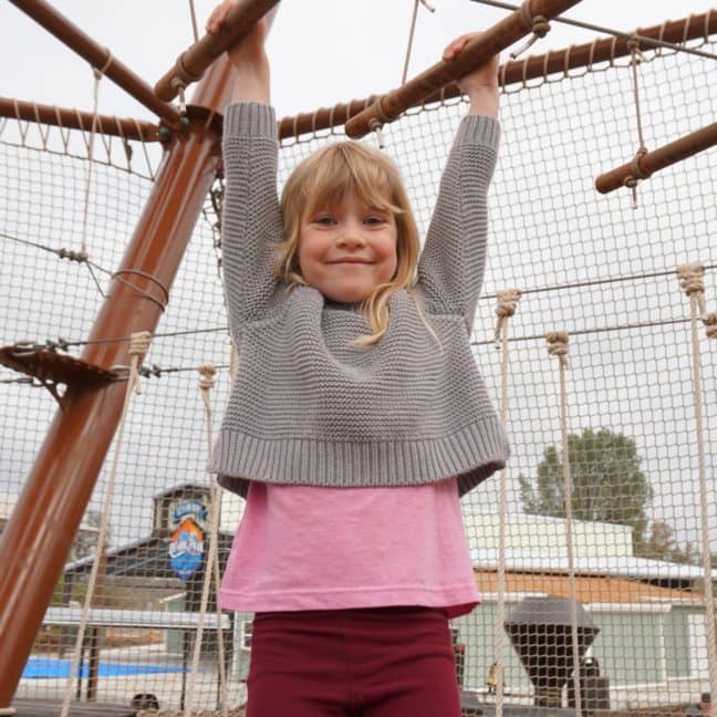 A young girl with blonde hair smiles at the camera while hanging from a rope structure at a playground, with other children in the background.