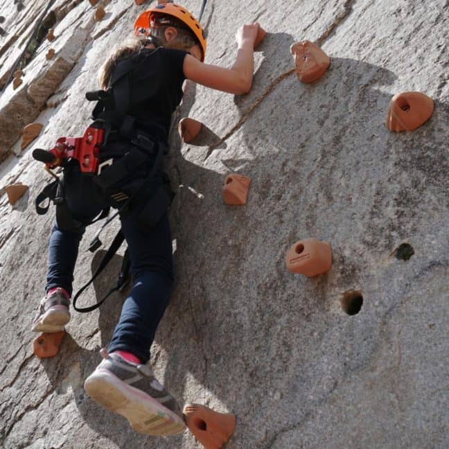 Two people wearing helmets and harnesses are climbing a grey artificial rock wall, focusing intently on their ascent.