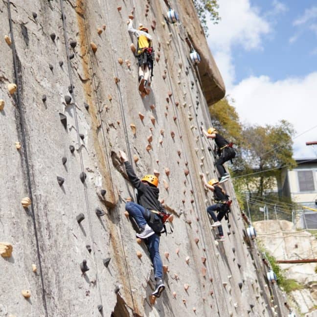 People climbing an outdoor artificial rock wall, equipped with safety harnesses and helmets, under a clear sky.