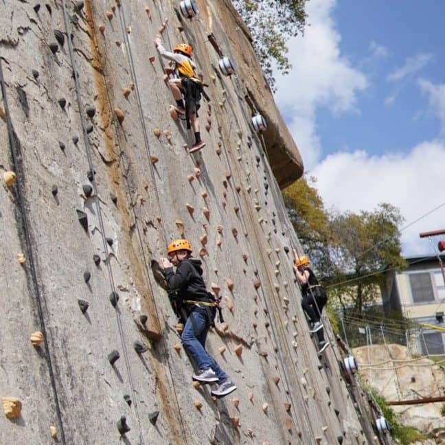 Three people wearing helmets and harnesses climb an outdoor artificial rock wall on a sunny day, with trees in the background.