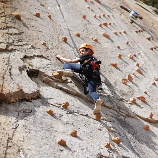 A woman wearing a helmet climbs an outdoor artificial rock wall, equipped with safety gear and ropes.