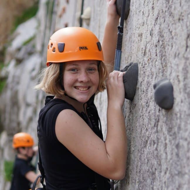 A young woman with a bright orange helmet climbing a rock wall, smiling at the camera, possibly taking advantage of a student discount on climbing gear.