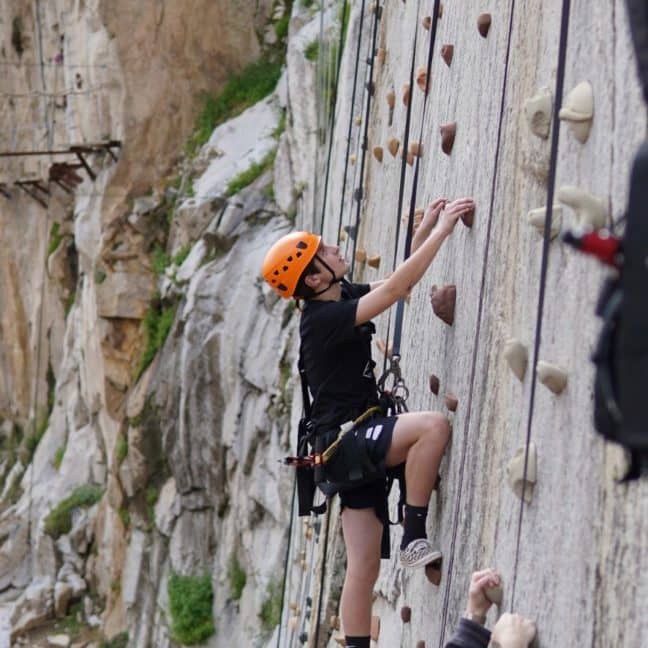 Person wearing a helmet and harness climbing an outdoor artificial rock wall, with others visible below.