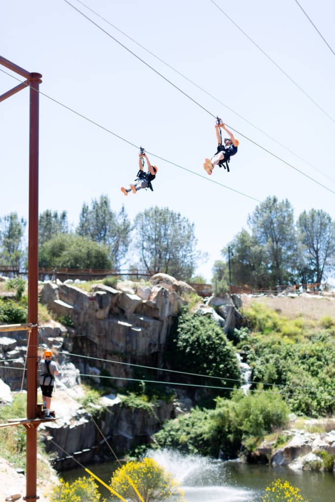 Two people zip-lining over a rocky landscape and water, dressed in outdoor sports gear, on a sunny day with a rafting package available.