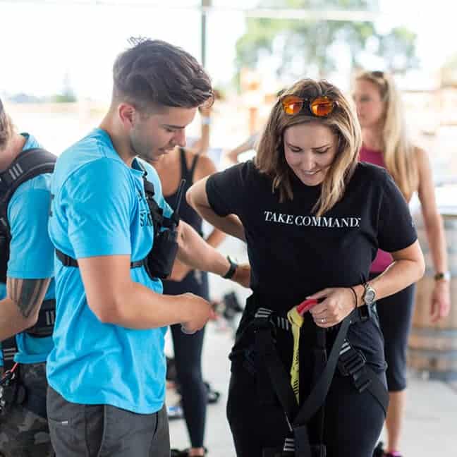 A woman and a man, both wearing harnesses, adjust their equipment before skydiving, as another person observes in the background.