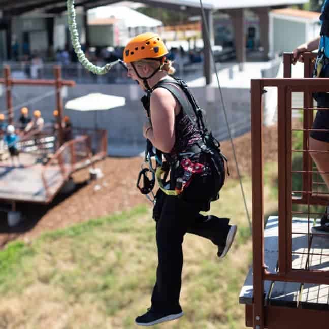 Person in orange helmet and harness zip-lining from a platform, with buildings and people in the background.