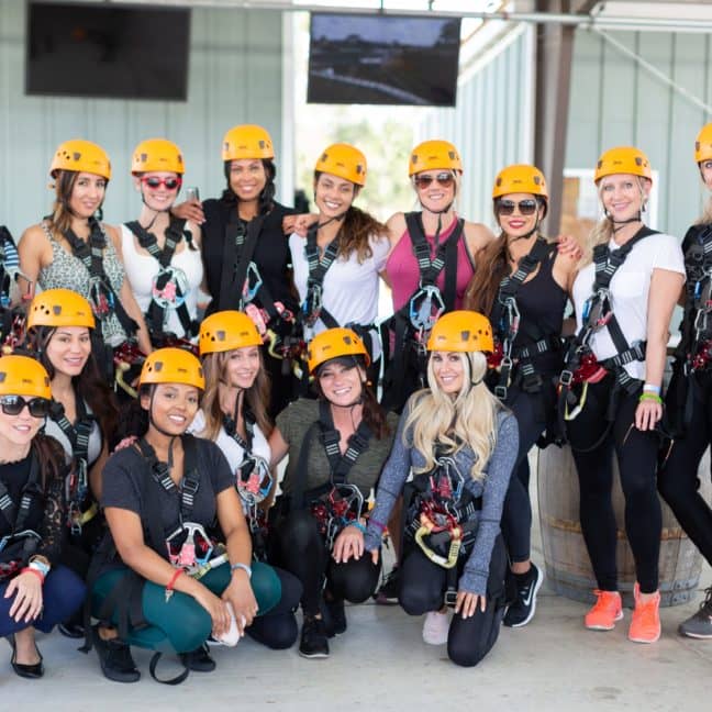 Group of women in helmets and harnesses posing together at an outdoor adventure facility.