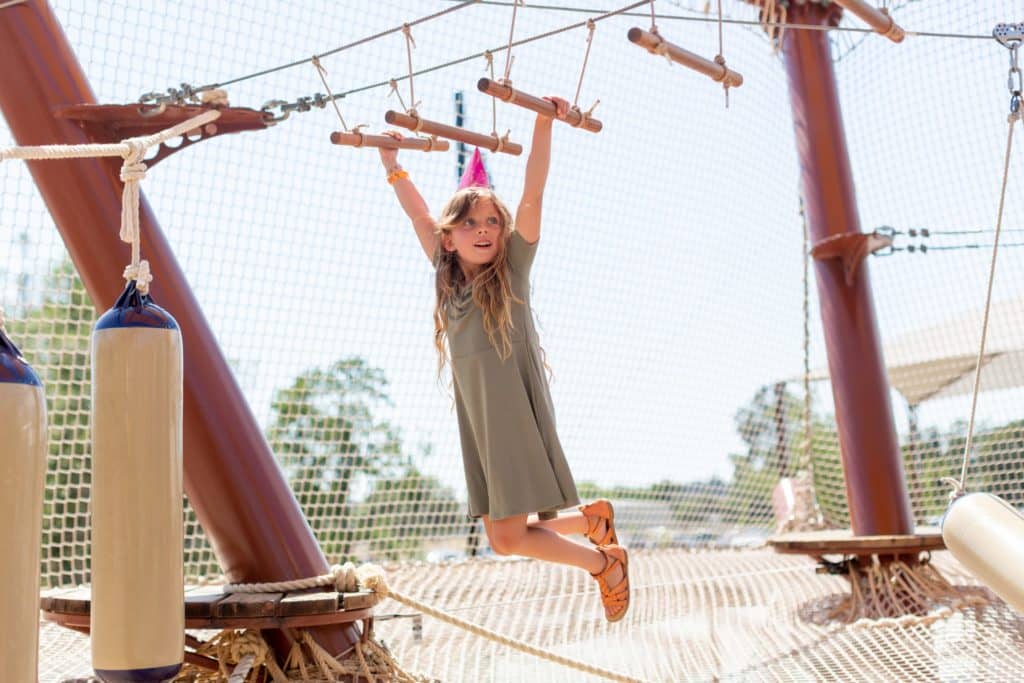 Young girl playing on a rope and wood adventure playground under a clear sky.