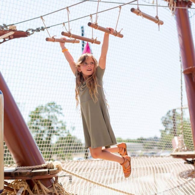 Young girl playing on a rope and wood adventure playground under a clear sky.