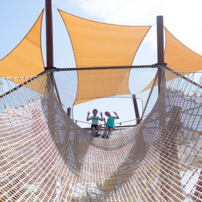 Two children playing on a large net playground under triangular sunshades on a sunny day.