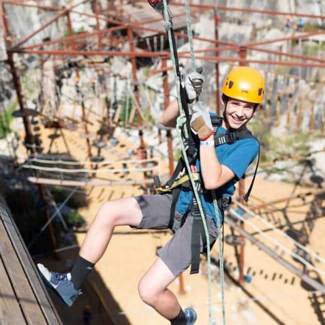 A student wearing a helmet and harness smiles while ziplining at an outdoor adventure park.