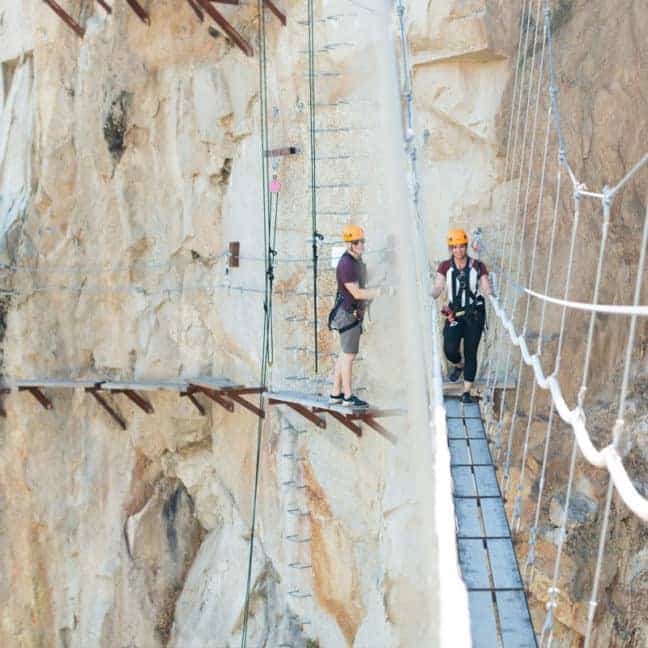 Two climbers in helmets crossing a narrow suspension bridge on a steep rocky cliff.