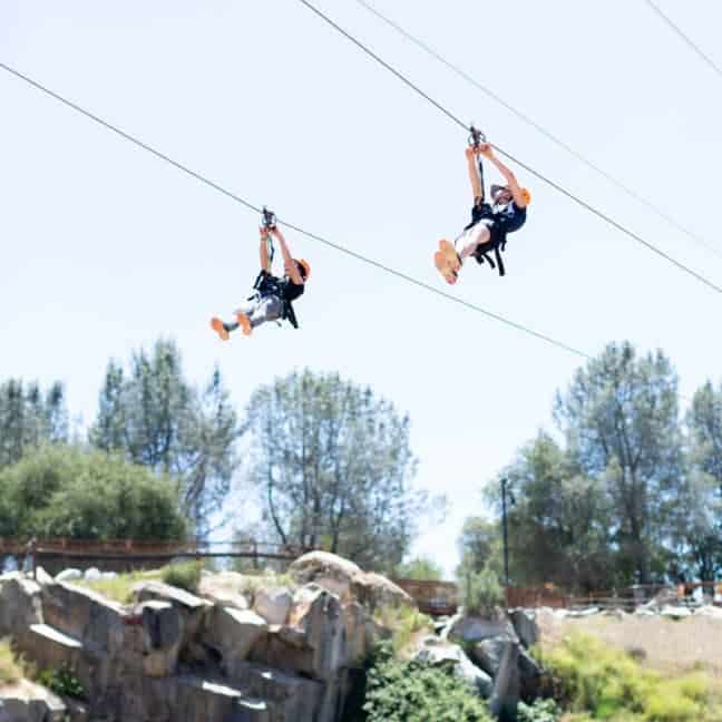 Two people ziplining over a rocky landscape with trees under a clear blue sky.