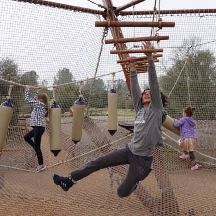 A child joyfully swings on a rope in a playground with others navigating hanging obstacles in the background during winter camps.