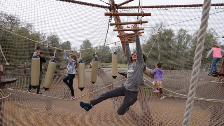 A child joyfully swings on a rope in a playground with others navigating hanging obstacles in the background during winter camps.