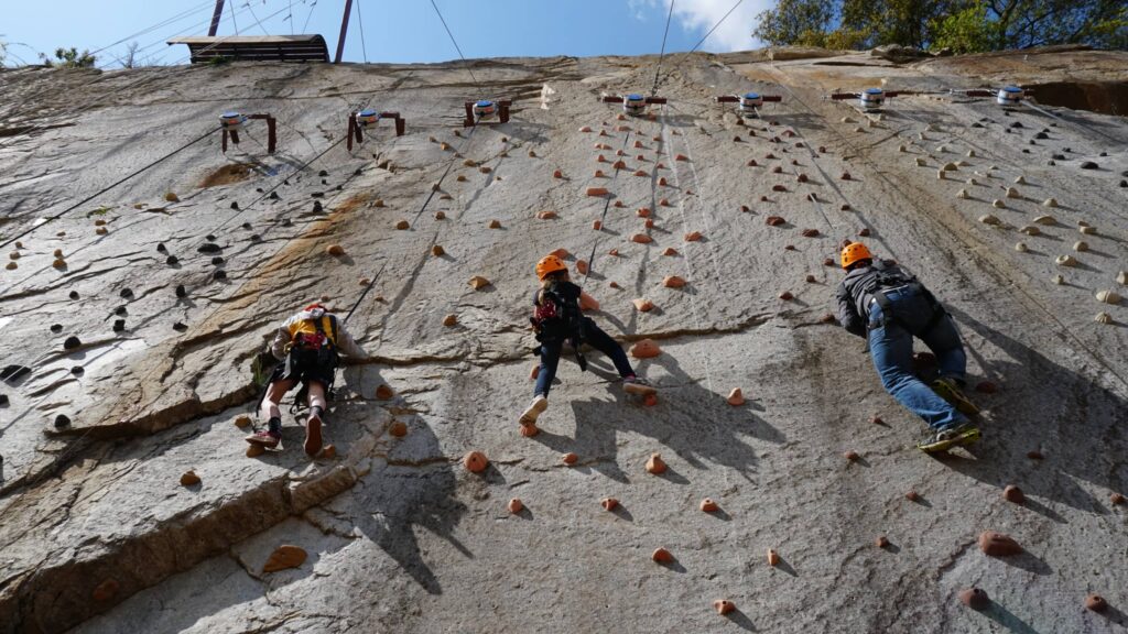 Three people with helmets and harnesses climb a large outdoor rock wall with scattered holds under a bright New Year sky.