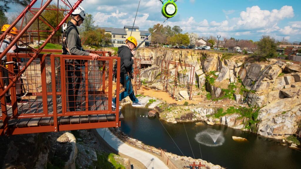 People wearing helmets prepare to zipline above a rocky quarry with a pond below, eager to kick off the New Year with adventurous spirits and big savings on thrills.