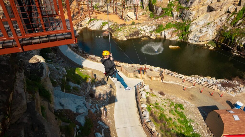 A person wearing a helmet and harness is zip-lining over a quarry with a pond and fountain below, embracing the thrill like finding big savings in the New Year.