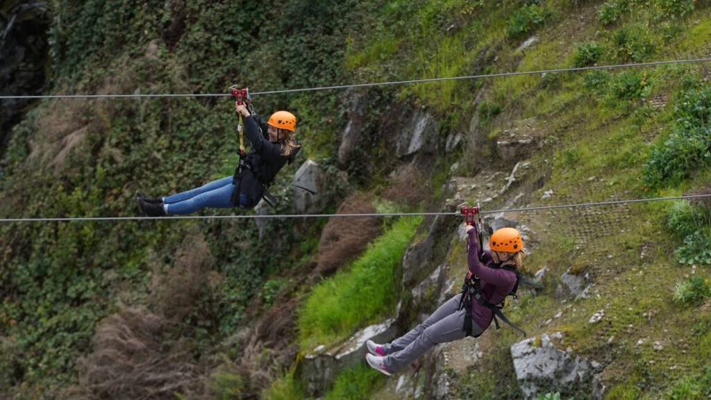 Two people, thrilled by the promise of big savings, zip line side by side over a lush, green, rocky landscape.