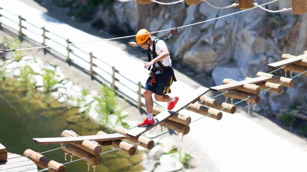 A person wearing a helmet and safety harness navigates a suspended wooden bridge over a scenic outdoor area, embracing the thrill of adventure with thoughts of big savings on New Year escapades.