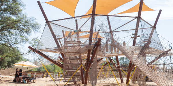 Large outdoor playground with climbing nets and orange shade sails, perfect for kids to enjoy while adults relax at picnic tables nearby—ideal for a Summer Birthday Party Sale celebration!.