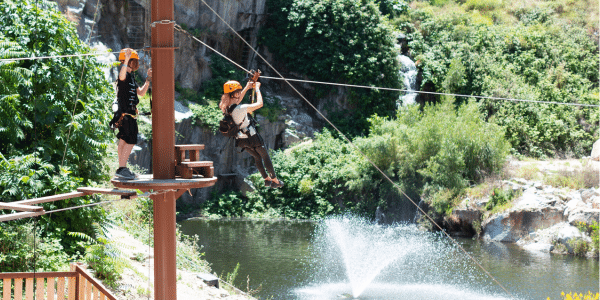 Two people wearing helmets zipline over a pond with a fountain, surrounded by greenery and rocky cliffs—perfect for adventurous fun at a Summer Birthday Party Sale!.