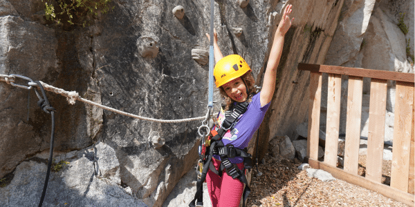 A young child wearing a helmet and harness stands in front of a rock climbing wall, smiling with arms raised, ready for an adventurous Summer Birthday Party.
