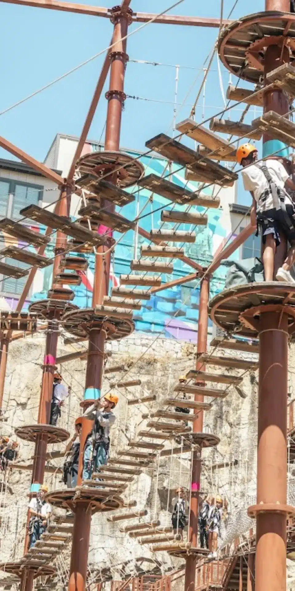 People wearing helmets and harnesses navigate a multi-level outdoor ropes course with wooden platforms and ropes, set against a rocky background and a building with a colorful mural—a true hero's challenge for 2025 adventure seekers.