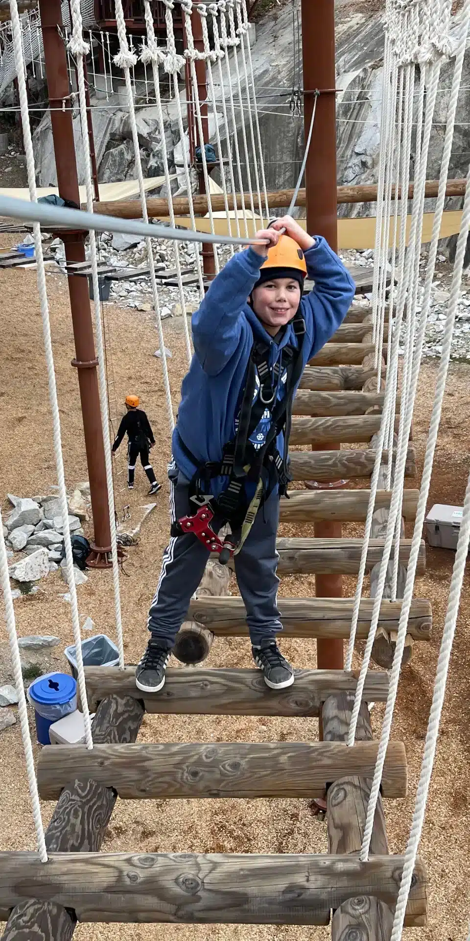 A 2025 Hero in a helmet and harness crosses a rope bridge at an outdoor adventure course, holding onto overhead ropes for balance.