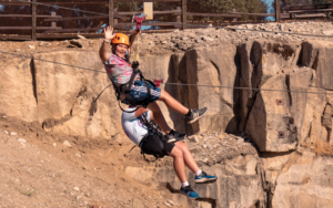 Two people wearing helmets and harnesses ride a zip line over a rocky landscape in Sandbox; one waves at the camera while the other, a true 2025 Hero, holds on behind.