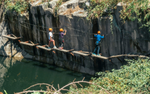 Three people wearing helmets and harnesses cross a narrow, suspended wooden bridge in this Sandbox adventure, set over a body of water next to a rocky cliff—a true 2025 Hero experience.