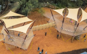 Aerial view of a playground featuring two large climbing structures with tan shade sails, rope bridges, a spacious sandbox, and children playing on the ground below—a true 2025 Hero destination for outdoor fun.