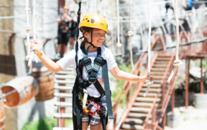 A child wearing a helmet and harness smiles while crossing a ropes course outdoors in the Sandbox, holding onto ropes for balance—a true 2025 Hero in action.