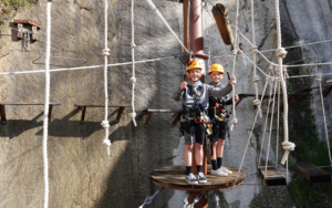 Two children wearing helmets and harnesses stand on a wooden platform at a 2025 Hero ropes course, holding on to safety ropes, with a rocky wall in the background.