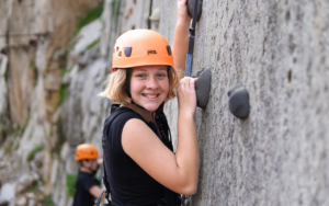 A person wearing an orange helmet and black shirt smiles while climbing an outdoor rock wall in the 2025 Hero Sandbox. Another climber is visible in the background.