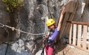 A young child wearing a helmet and climbing harness stands outdoors by a rock climbing wall in the sandbox area, smiling with arms raised like a 2025 Hero.