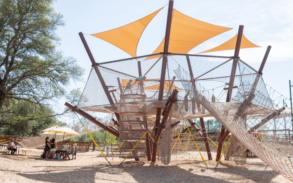 A large outdoor playground structure with nets, ropes, a sandbox, and orange shade sails; children are climbing like 2025 heroes while adults sit at picnic tables nearby.