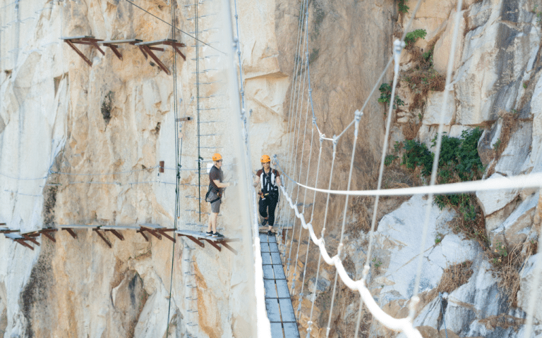 Two people wearing helmets and harnesses stand on a suspended rope bridge attached to a rocky cliffside, their safety equipment visible as they embrace the thrill of this 2025 Hero adventure in nature’s own sandbox.