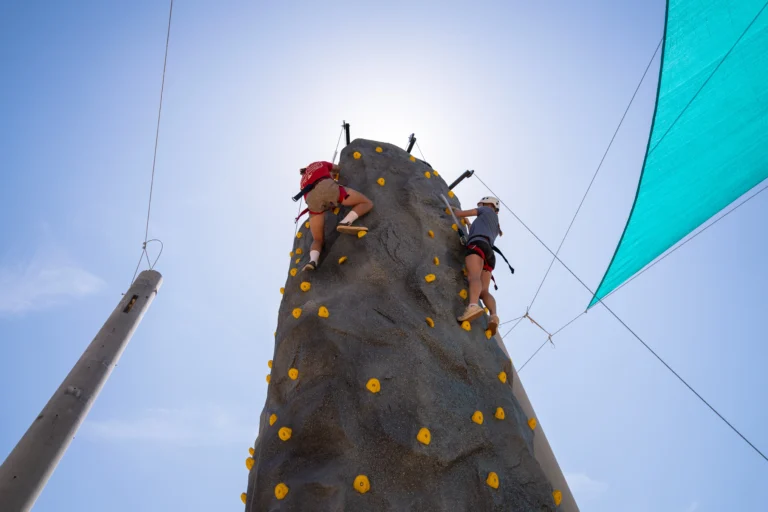 Two people wearing harnesses climb an outdoor artificial rock wall under a sunny sky, with safety ropes and sunshades visible.