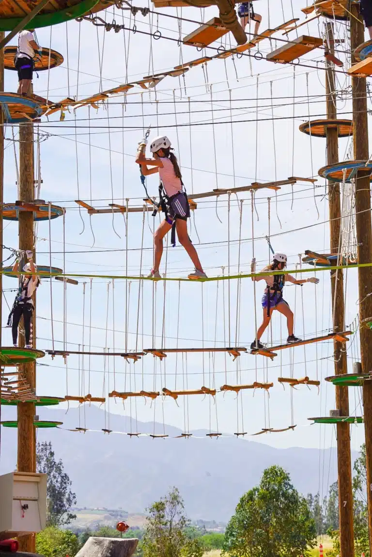 People wearing helmets and harnesses navigate a multi-level outdoor ropes course, balancing on wooden planks and ropes with a mountainous landscape in the background.